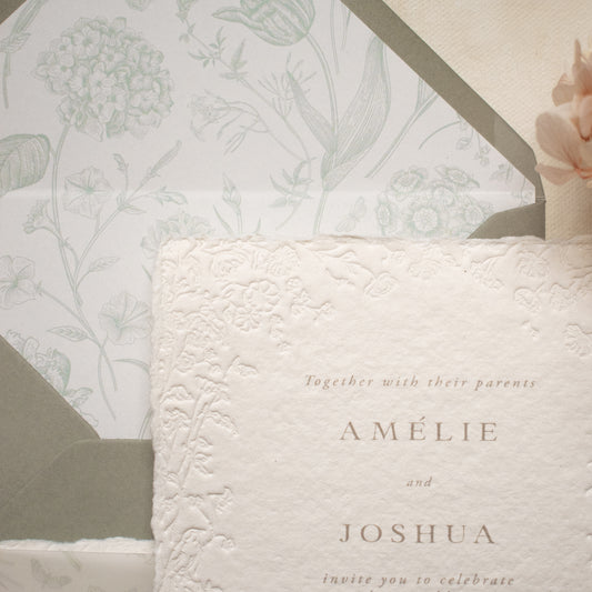 A close-up of a wedding invitation card with embossed floral detailing, layered over a matching floral-patterned envelope liner and a dusty sage envelope.