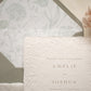A close-up of a wedding invitation card with embossed floral detailing, layered over a matching floral-patterned envelope liner and a dusty sage envelope.