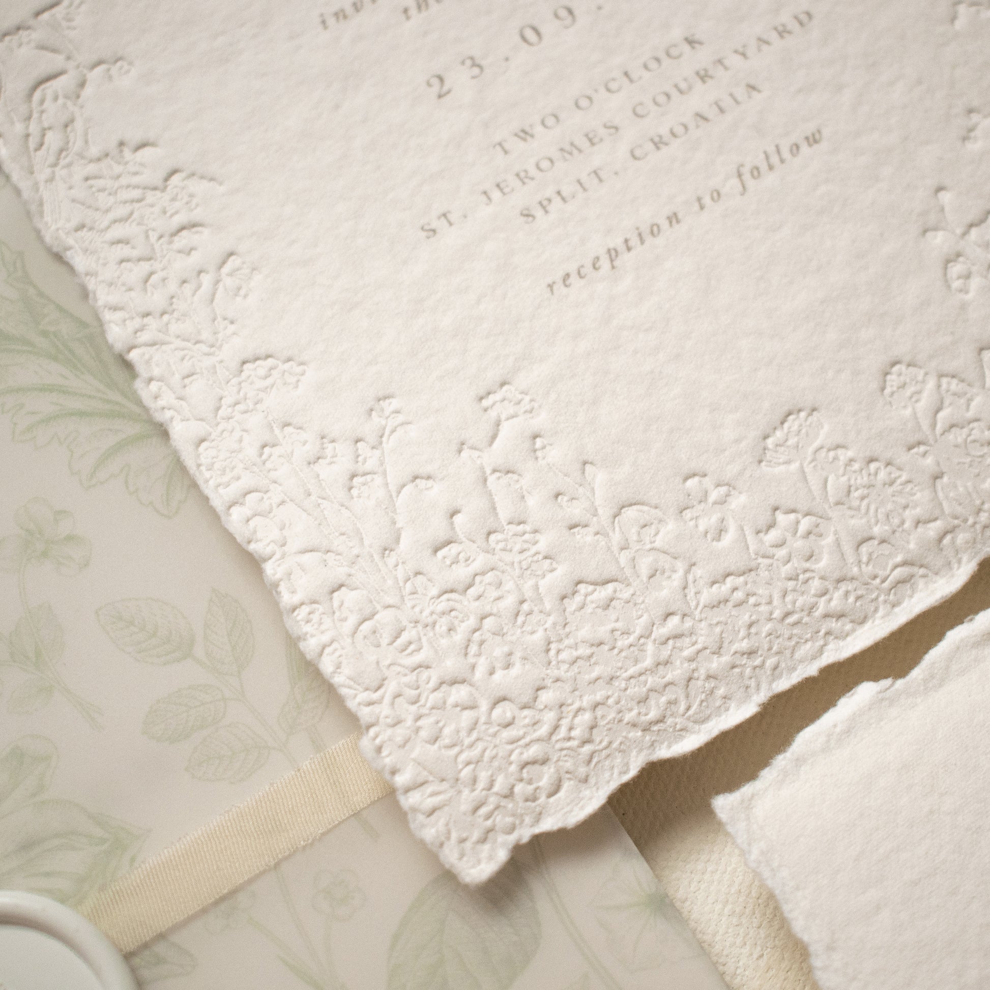 A close-up of a white textured paper invitation card with blind letterpress floral borders, layered over a dusty sage floral vellum wrap