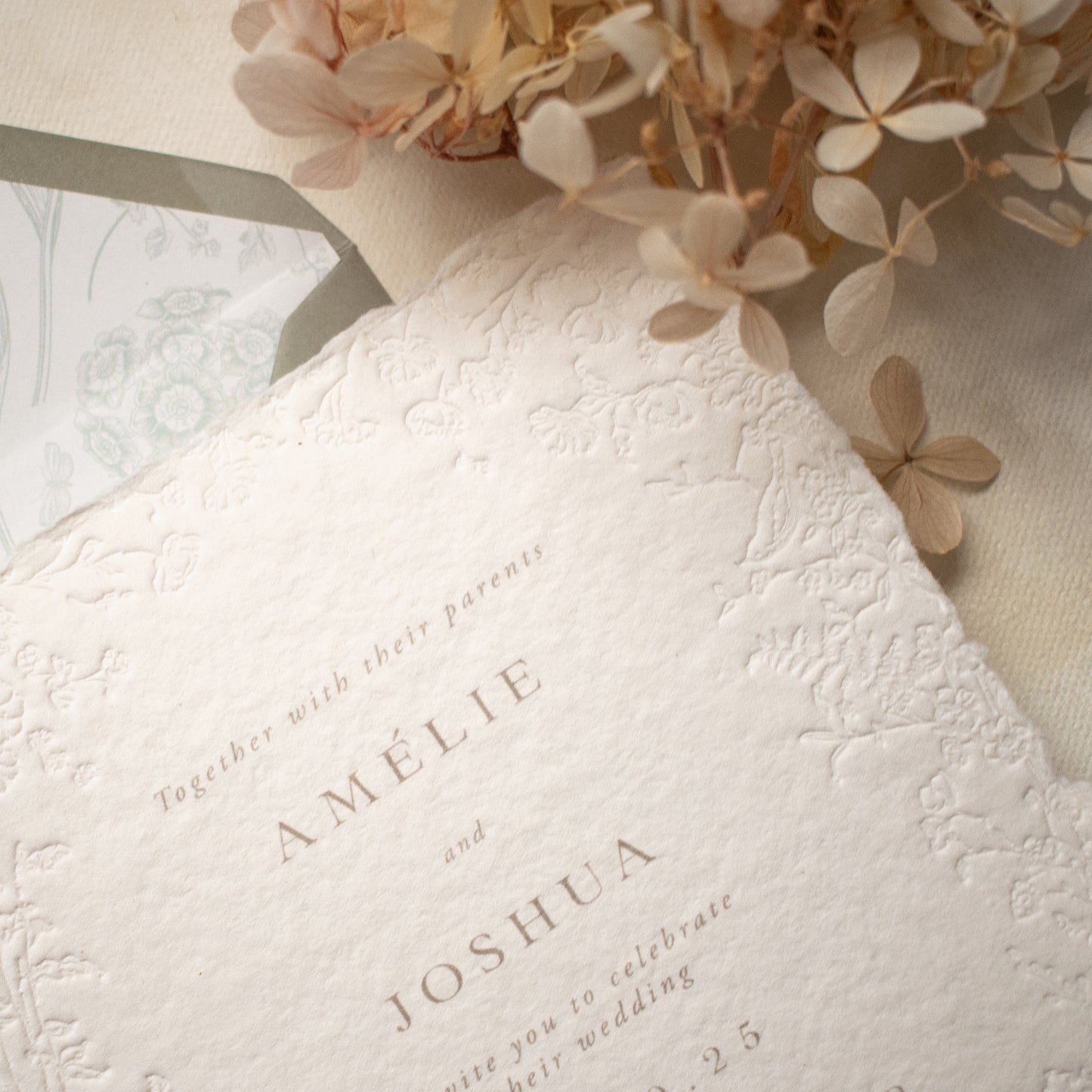 A close-up of a white textured paper invitation card with blind letterpress floral borders, layered over a dusty sage floral envelope