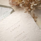 A close-up of a white textured paper invitation card with blind letterpress floral borders, layered over a dusty sage floral envelope