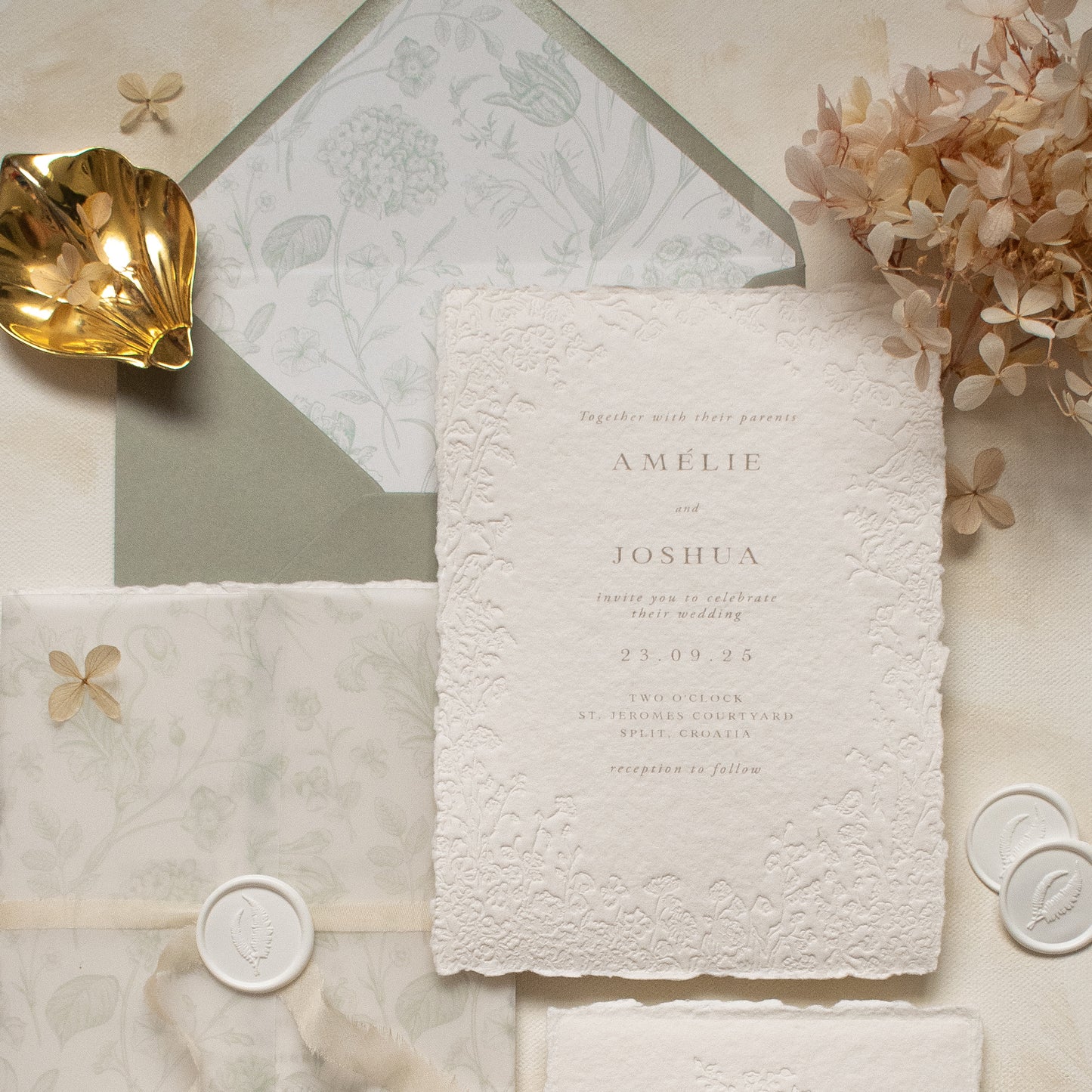 A close-up of a white textured paper invitation card with blind letterpress floral borders, layered over a dusty sage floral vellum wrap and matching sage green envelope with floral printed liner, styled with dried flowers and white wax seals