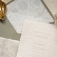 A close-up of a white textured paper invitation card with blind letterpress floral borders, layered over a dusty sage floral envelope