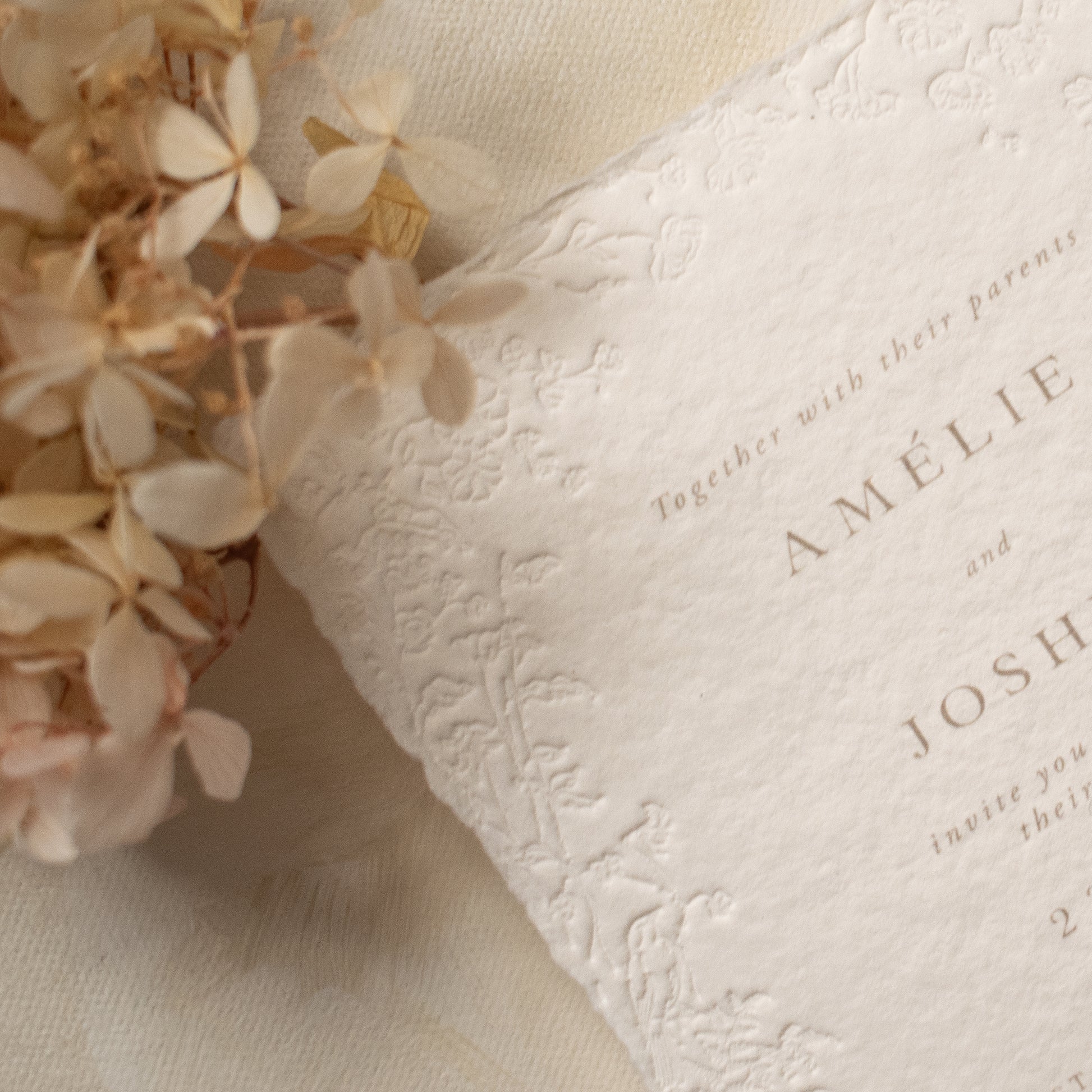 A close-up of a white textured paper invitation card with blind letterpress floral borders, styled with beige dried flowers